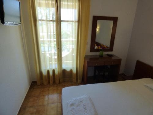 A cozy hotel room with tiled floors, yellow curtains framing a window view, a wooden desk with a mirror, and neatly arranged bedding invites travelers seeking comfort and simplicity.