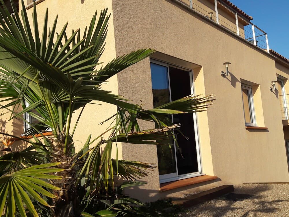 A serene vacation rental features a modern beige exterior with large windows, a balcony, and lush palm foliage, ideal for travelers seeking a tranquil tropical retreat.
