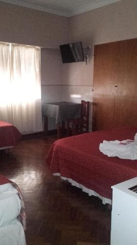 A cozy hotel room with wooden floors, red bedding, a small dining area, and a wall-mounted TV offers a comfortable stay for travelers seeking simplicity and functionality.