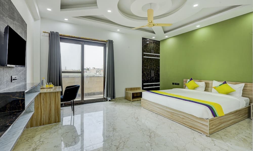 A cozy hotel room with a vibrant green accent wall, marble flooring, a neatly made bed with colorful pillows, a modern desk setup, and a balcony view, ideal for travelers seeking comfort and style.