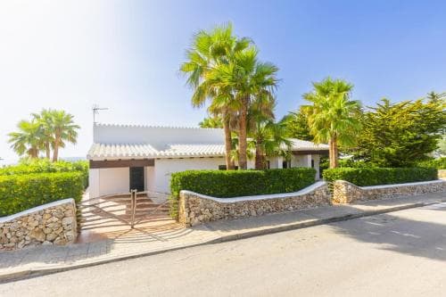 A serene Mediterranean villa entrance framed by lush palm trees and manicured hedges under a clear blue sky, ideal for a tranquil vacation retreat.