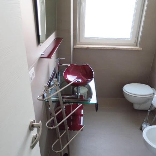A modern bathroom features a sleek red sink, white toilet, and natural light from a window, creating a clean and functional space ideal for travelers seeking comfort in accommodations.