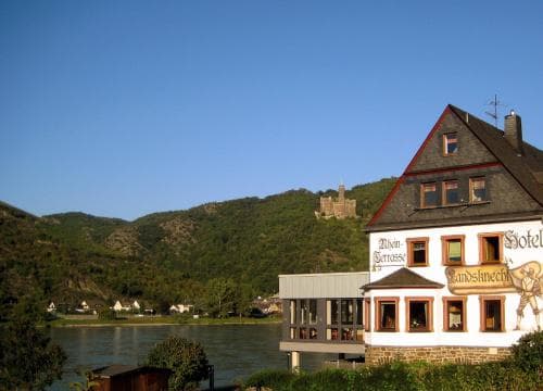 A charming riverside hotel with traditional architecture overlooks a serene river valley under a clear blue sky, ideal for travelers seeking a peaceful retreat in a picturesque European setting.