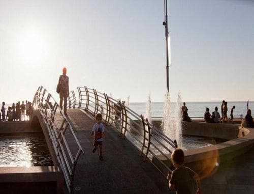 A vibrant seaside promenade features a modern bridge with playful fountains, bustling with families enjoying the sunny day, ideal for family-friendly beachside getaways.