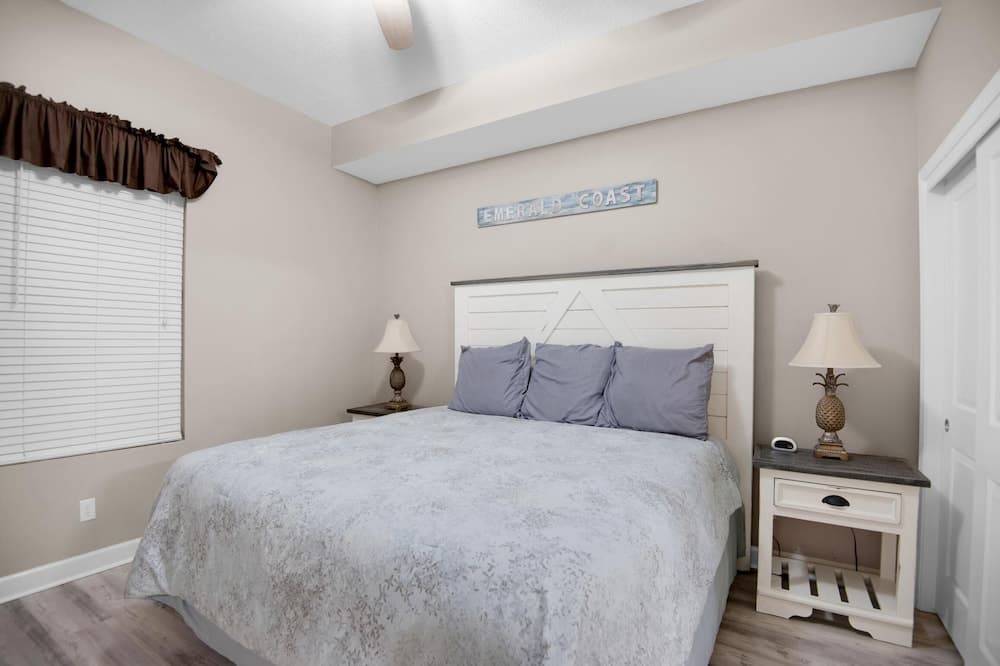 A cozy bedroom with a white bed adorned by gray linens, pineapple lamps on nightstands, and an "Emerald Coast" sign above the headboard, ideal for travelers seeking a serene coastal retreat.