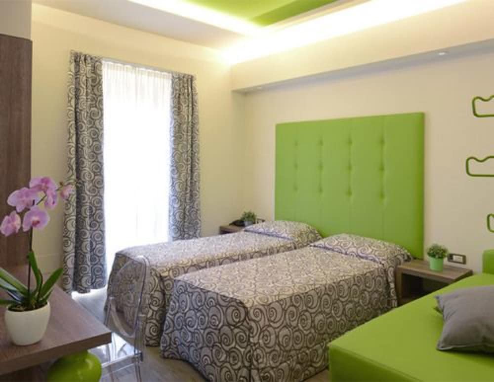 A cozy hotel room features two neatly made beds with patterned linens, a vibrant green headboard, and soft natural light filtering through patterned curtains, creating a welcoming atmosphere ideal for travelers seeking comfort and relaxation.