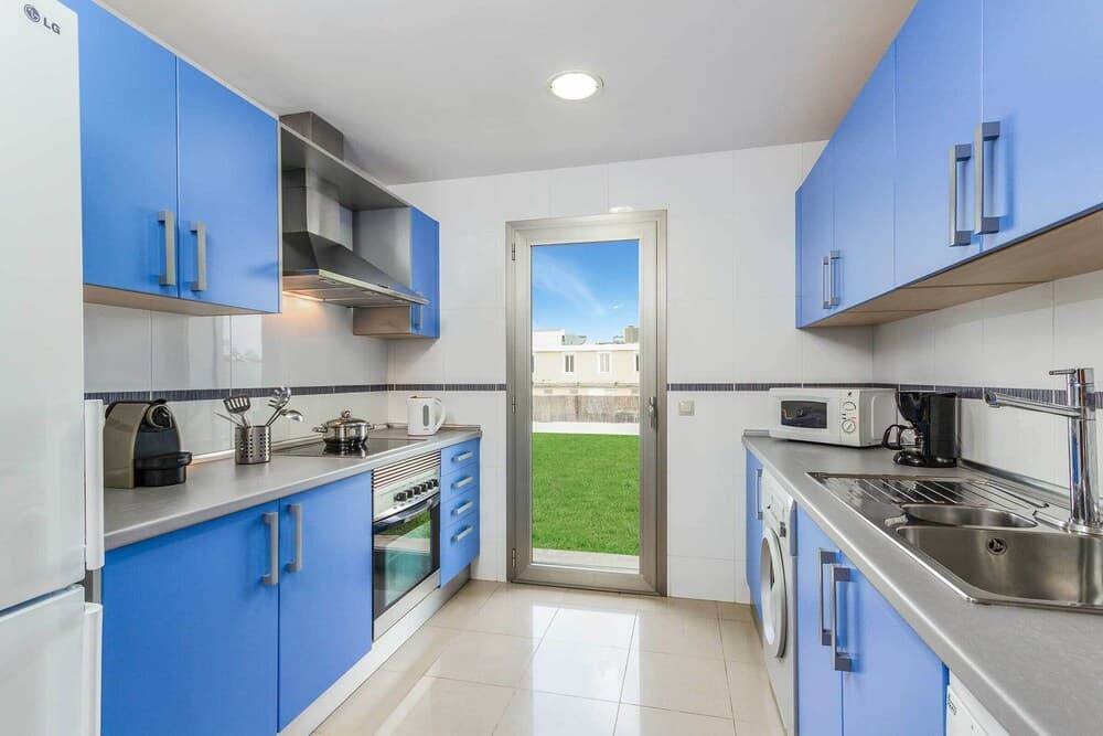 A modern kitchen with vibrant blue cabinets and stainless steel appliances opens to a serene outdoor view through a glass door, ideal for travelers seeking a cozy and functional rental space.