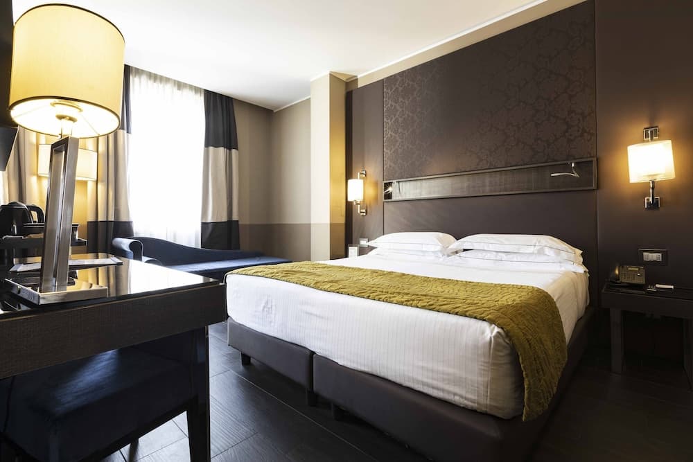 A cozy hotel room features a neatly made bed with white linens and a green throw, warm lighting from bedside lamps, and a stylish desk area, ideal for travelers seeking comfort and functionality.