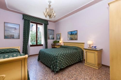 A cozy hotel room with twin beds adorned in green patterned bedding, framed by elegant curtains and a chandelier, evokes a serene and inviting atmosphere ideal for travelers seeking comfort and charm.