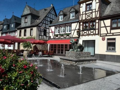 A charming European village square features half-timbered buildings, a playful cat fountain, and vibrant red umbrellas over outdoor seating, ideal for travelers seeking a quaint and relaxing atmosphere.