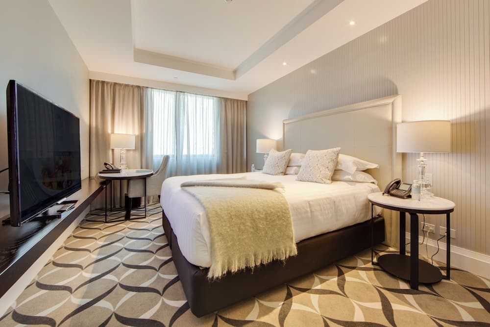 A cozy hotel room features a neatly made bed with plush pillows, a stylish patterned carpet, modern bedside tables with lamps, a desk by the window, and a flat-screen TV, ideal for travelers seeking comfort and relaxation.