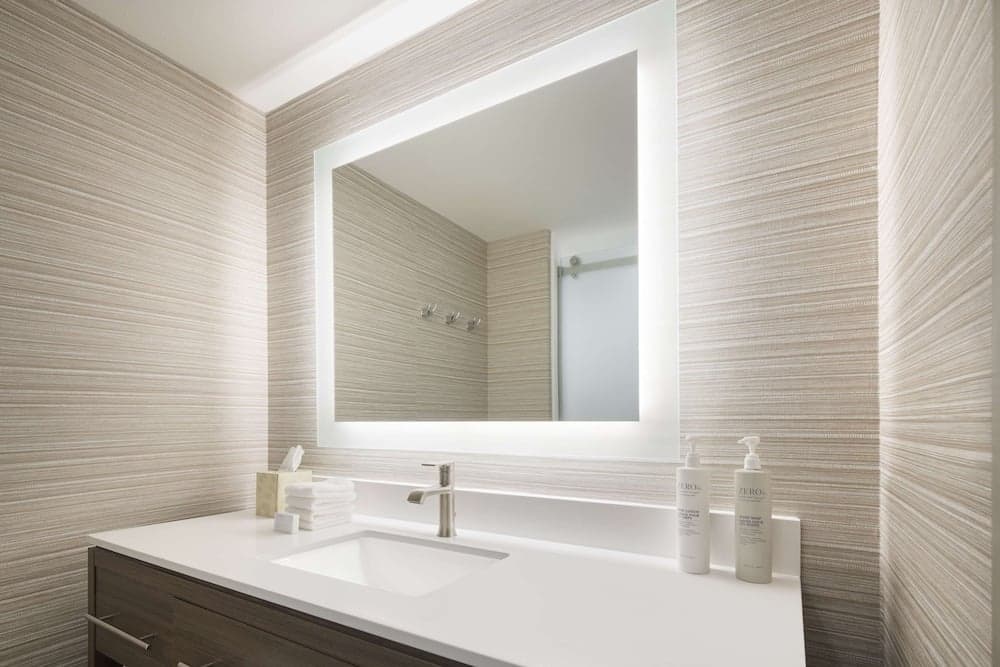 A serene hotel bathroom features a modern vanity with a sleek sink, illuminated mirror, striped wallpaper, and neatly arranged toiletries, ideal for travelers seeking comfort and style.