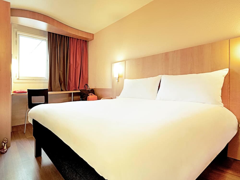 A cozy hotel room features a neatly made bed with white linens, wooden furnishings, soft lighting, and a window with colorful curtains, ideal for travelers seeking comfort and relaxation.