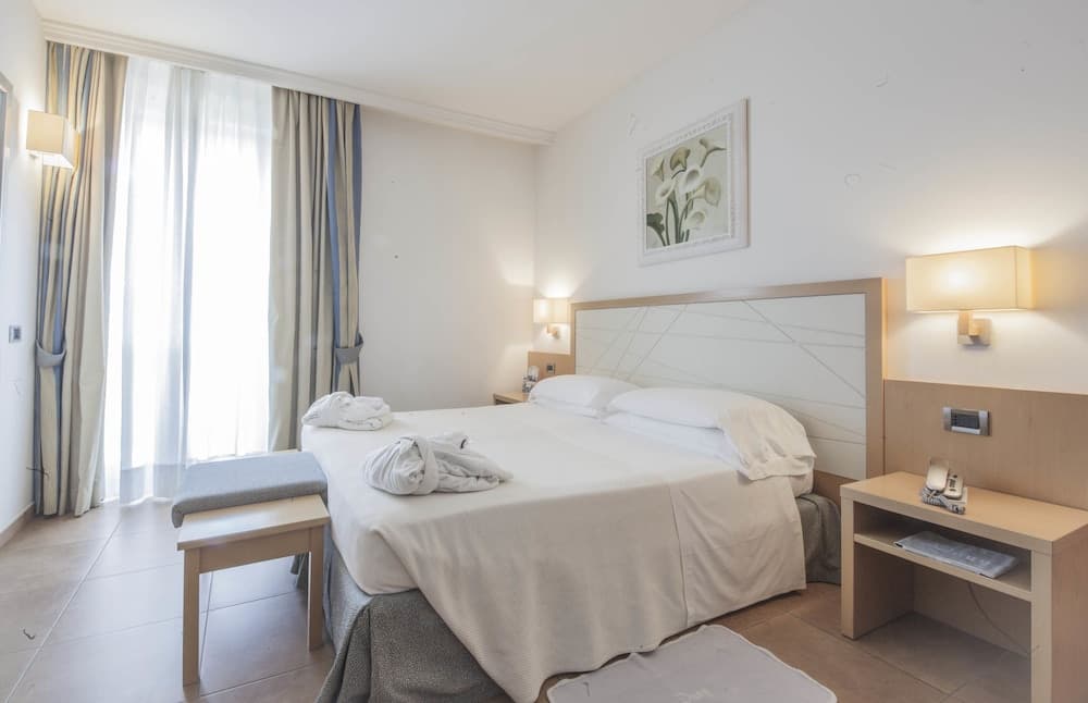 A cozy hotel room with a neatly made bed, soft lighting from wall-mounted lamps, and a serene view through sheer curtains, ideal for travelers seeking comfort and relaxation.