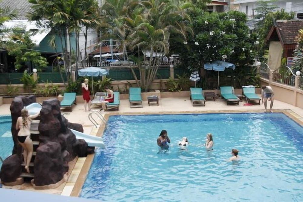 A vibrant tropical resort pool scene features guests relaxing on lounge chairs under umbrellas and enjoying the water near a rock slide, ideal for vacationers seeking leisure and relaxation in a sunny, palm-fringed setting.