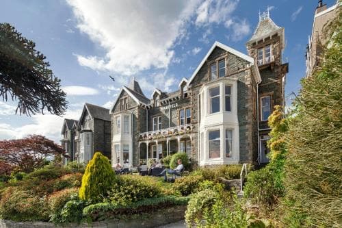 A charming Victorian-style hotel with lush gardens and guests relaxing outdoors under a bright blue sky, ideal for travelers seeking a cozy retreat in a picturesque setting.