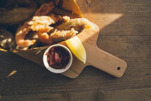 A rustic wooden table sets the stage for a cozy dining experience featuring a golden fish burger, crispy fries, a lemon wedge, and a small bowl of tangy sauce, perfect for travelers seeking a casual yet flavorful meal.
