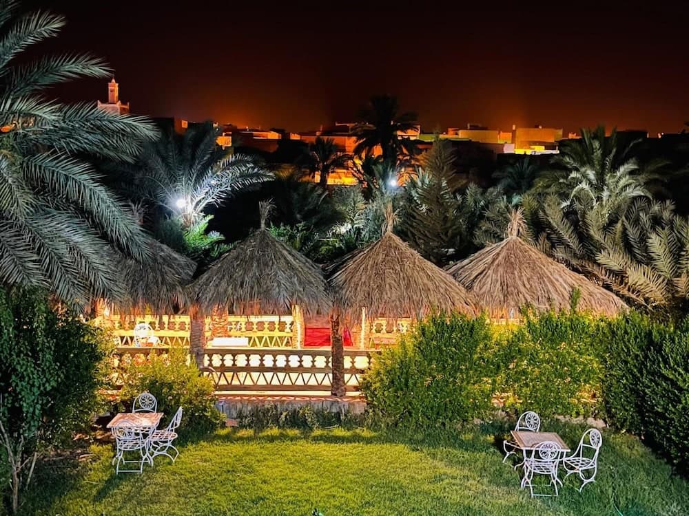 A serene night scene at an exotic resort features thatched-roof huts surrounded by lush palm trees and greenery, illuminated by warm lights, creating a tranquil atmosphere ideal for romantic getaways or peaceful retreats.