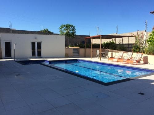 A serene private villa poolside scene features a clear blue pool, sun loungers, and lush greenery under a bright sky, ideal for relaxing summer vacations.