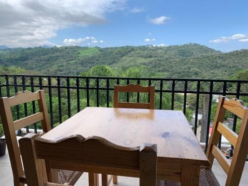 A serene balcony with a wooden table and chairs overlooks lush green hills under a partly cloudy sky, ideal for travelers seeking a peaceful retreat in a mountainous region.