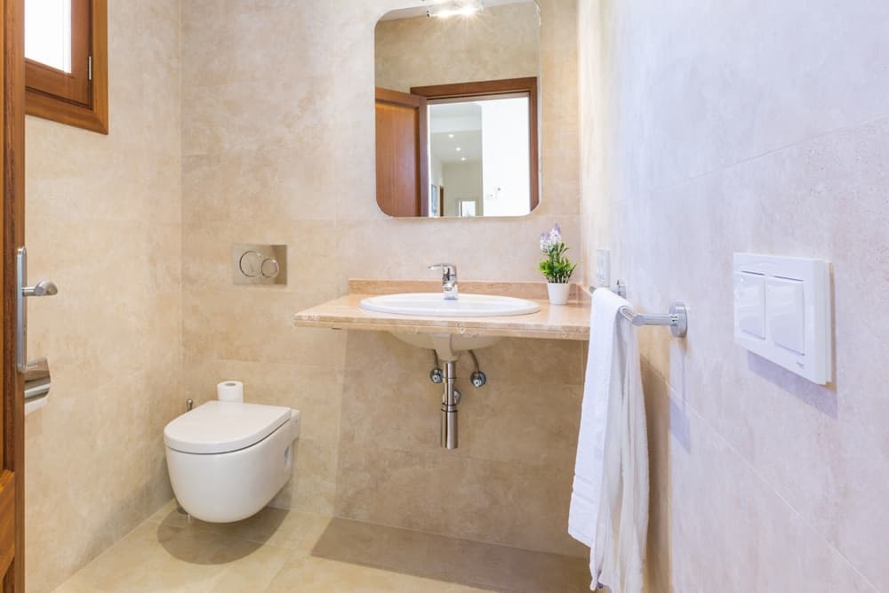 A serene and modern bathroom features beige tiled walls, a sleek white toilet, a countertop sink with a mirror, a small potted plant, and a neatly hung towel, ideal for travelers seeking comfort and cleanliness in their accommodations.