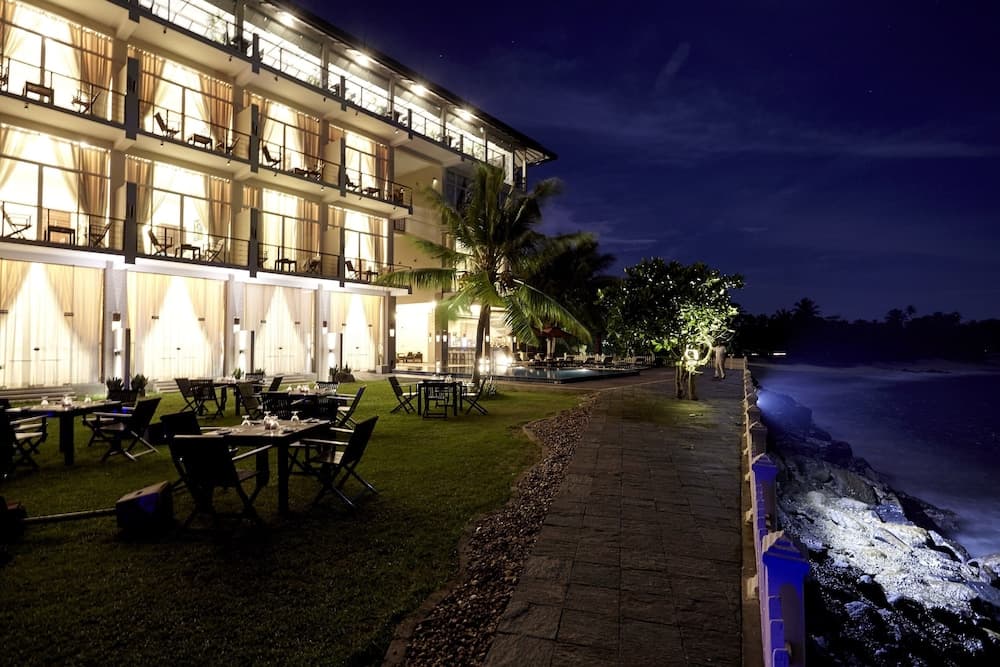 A serene coastal resort at night features illuminated balconies, outdoor dining tables, palm trees, and a stone pathway leading to the ocean, ideal for romantic getaways or luxurious beachfront stays.