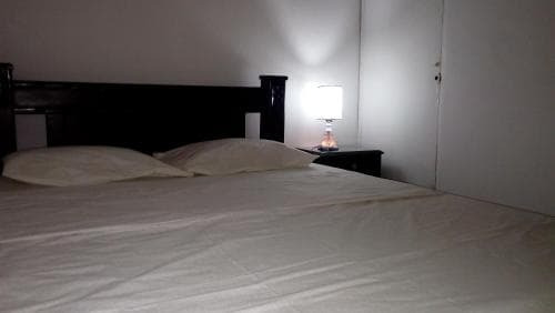 A cozy hotel room features a neatly made bed with white linens, a dark wooden headboard, and a softly glowing bedside lamp, creating a serene atmosphere ideal for travelers seeking restful accommodations.