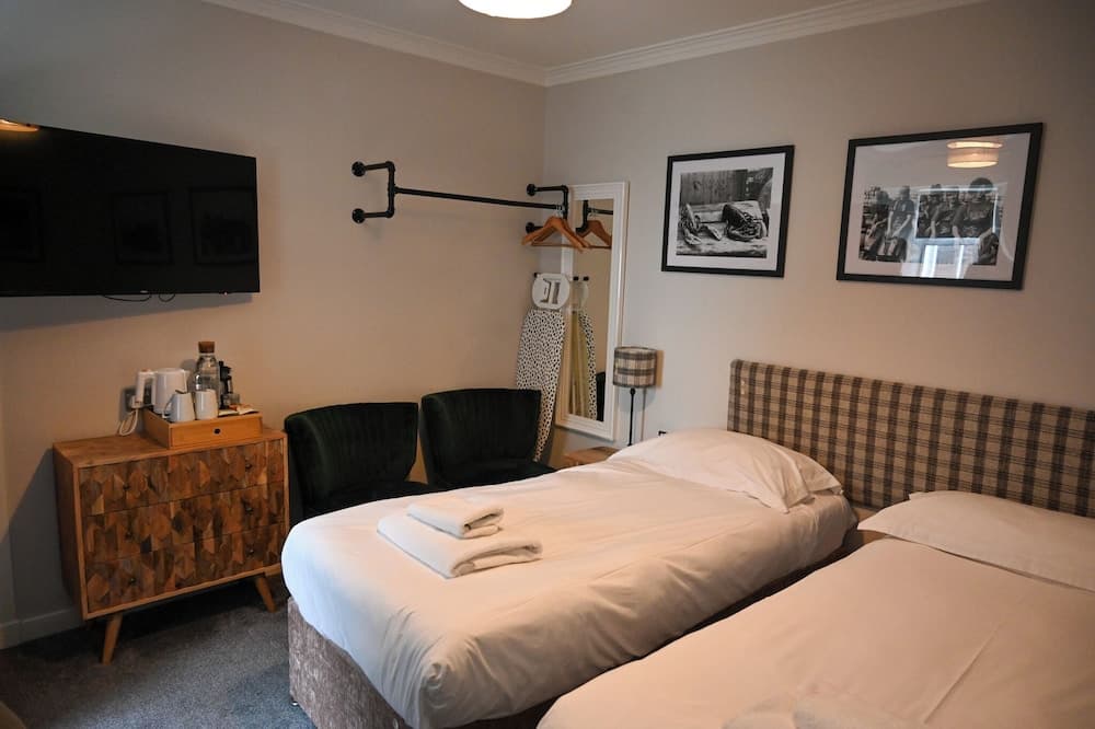 A cozy hotel room features two neatly made beds, modern furnishings including a patterned dresser and green armchairs, framed black-and-white artwork on the walls, and a mounted TV, ideal for travelers seeking comfort and style.