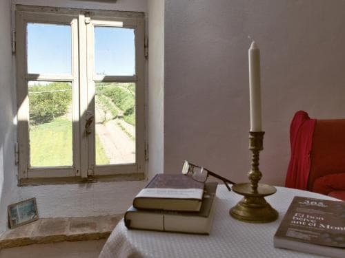 A serene indoor retreat features a rustic window overlooking a tranquil countryside path, paired with a cozy table adorned with books, a candlestick, and glasses, ideal for a peaceful reading getaway.