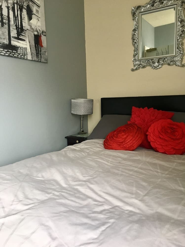 A cozy bedroom scene features a neatly made bed adorned with vibrant red floral pillows, a stylish bedside lamp, an ornate mirror reflecting the room's charm, and a black-and-white wall art piece, ideal for travelers seeking a comfortable and aesthetically pleasing accommodation.