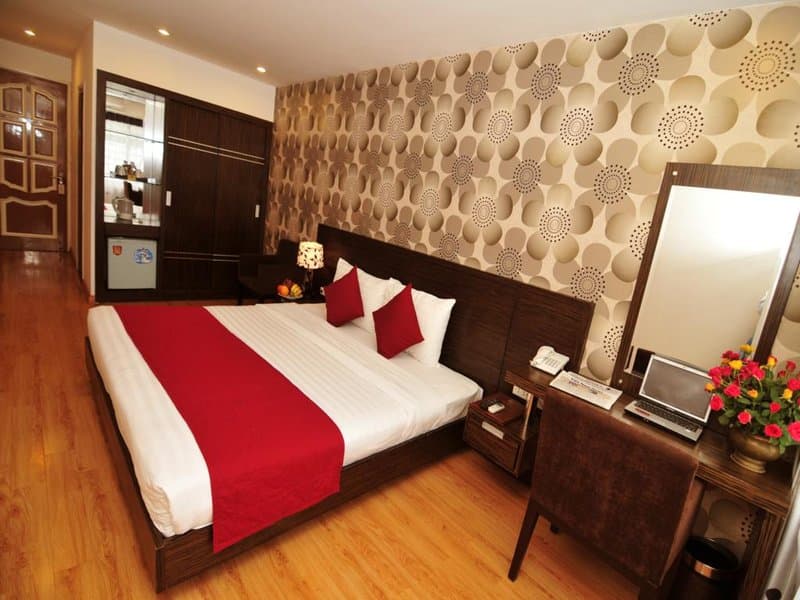 A cozy hotel room features a neatly made bed with red accents, a stylish patterned wall, and a desk with vibrant flowers, ideal for travelers seeking comfort and functionality.