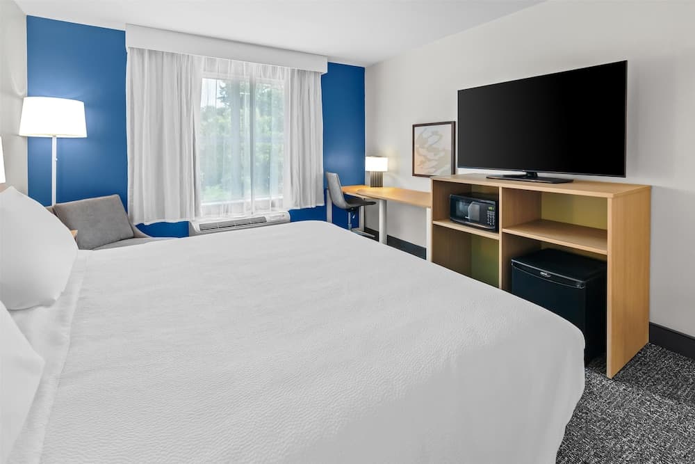 A cozy hotel room features a neatly made bed, a desk by the window, and a wooden entertainment unit with a TV, microwave, and mini-fridge, ideal for travelers seeking comfort and convenience.