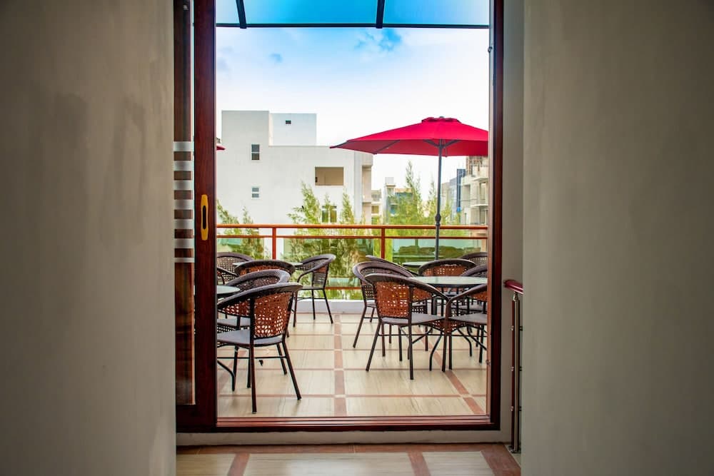 A serene balcony with wicker furniture under a red umbrella invites relaxation, framed by an open door leading to a modern urban view, perfect for travelers seeking a cozy retreat.