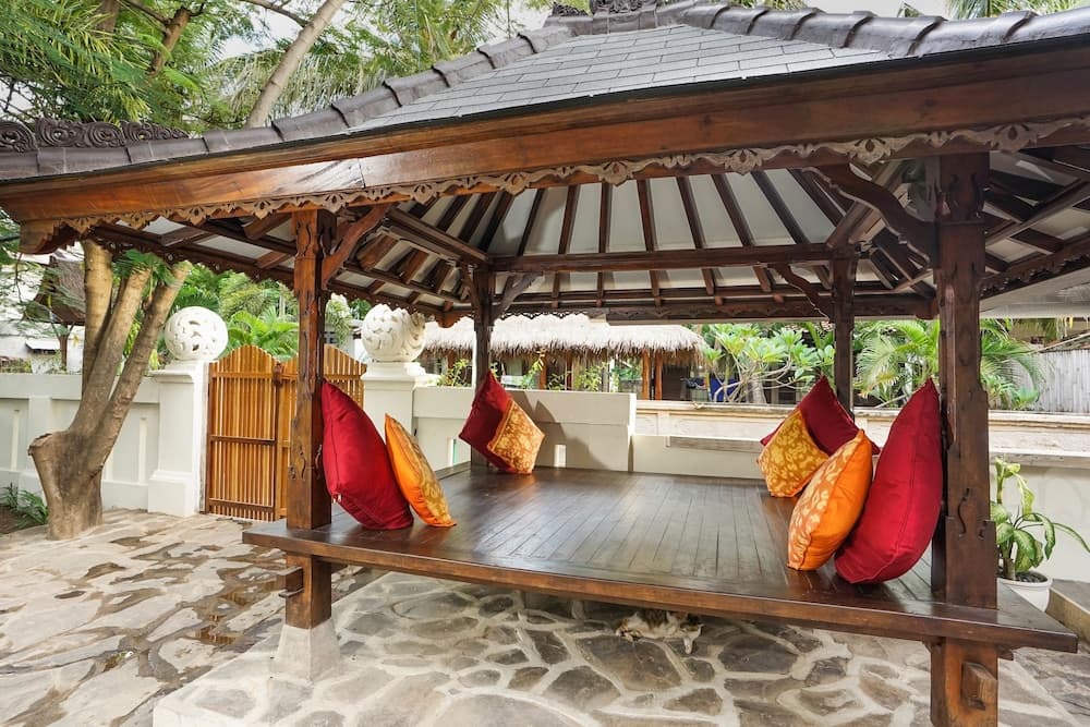 A serene tropical retreat features a wooden pavilion adorned with vibrant red and orange cushions, set on a stone-paved patio surrounded by lush greenery, ideal for relaxing vacations or cultural exploration.