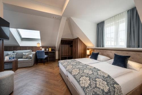 A cozy hotel room with a sloped ceiling, skylight, and large window offers a serene atmosphere, featuring a neatly made bed with floral bedding, modern furnishings, and warm lighting, ideal for travelers seeking comfort and relaxation.