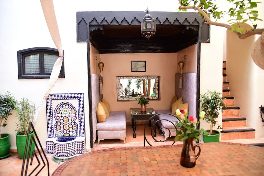 A serene Moroccan-style courtyard features intricate tilework, plush seating under a carved archway, vibrant greenery, and warm lighting, ideal for travelers seeking a tranquil cultural retreat.