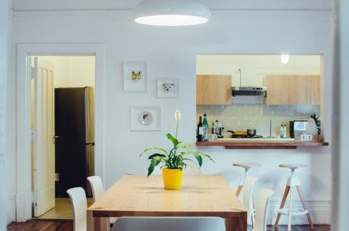 A cozy, modern kitchen-dining area with white walls, a wooden table adorned with a vibrant yellow potted plant, and a well-equipped kitchen in the background, ideal for travelers seeking a comfortable self-catering stay.