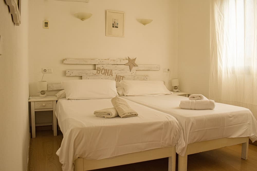 A cozy hotel bedroom with twin beds adorned with neatly folded towels, a rustic wooden headboard inscribed "BONA NIT," soft lighting, and sheer curtains filtering natural light, ideal for travelers seeking a serene and inviting accommodation.
