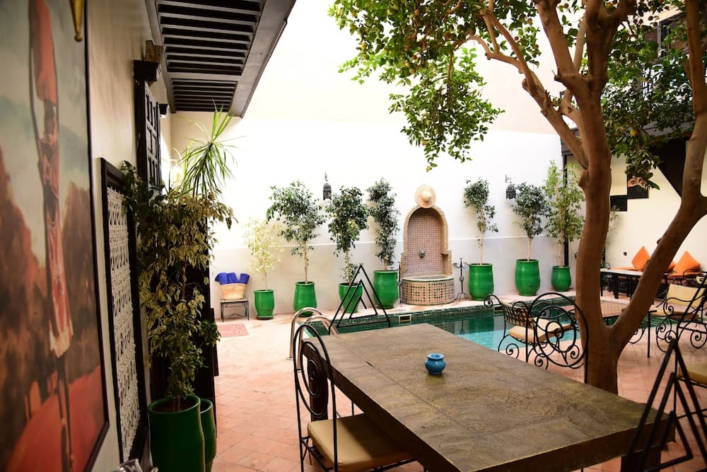A serene courtyard with a tiled fountain, green potted plants, and a shaded dining area offers a tranquil oasis for travelers seeking relaxation in a traditional setting.