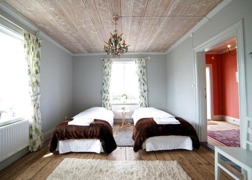 A cozy guesthouse room features twin beds with brown throws, wooden floors, a chandelier, and floral curtains, creating a warm and inviting atmosphere ideal for travelers seeking comfort and charm.