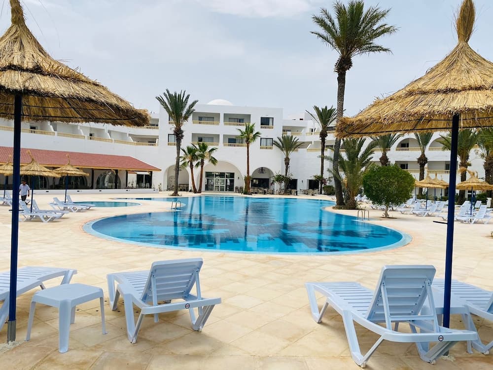 A serene tropical resort features a sparkling pool surrounded by palm trees, thatched umbrellas, and lounge chairs, ideal for relaxing vacations in a sunny destination.