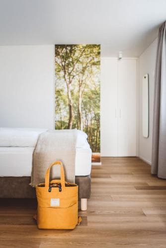 A cozy hotel room features a serene forest mural, minimalist decor, and a vibrant yellow tote bag on wooden flooring, ideal for travelers seeking relaxation and simplicity.