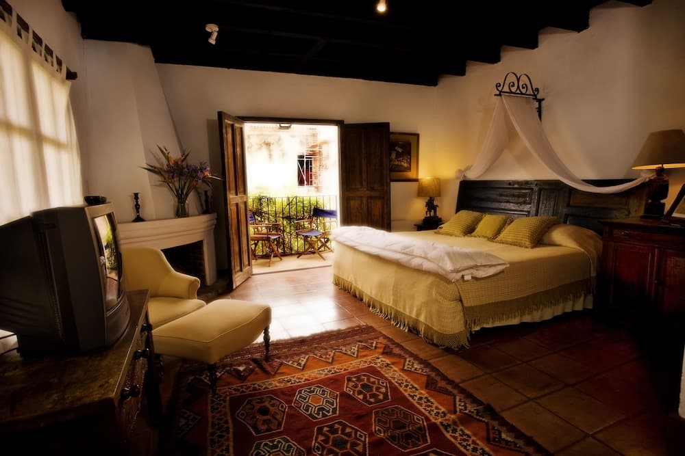 A cozy bedroom with rustic charm features a canopy bed, vibrant rug, wooden beams, and an open door leading to a balcony, ideal for travelers seeking a serene retreat in a traditional setting.
