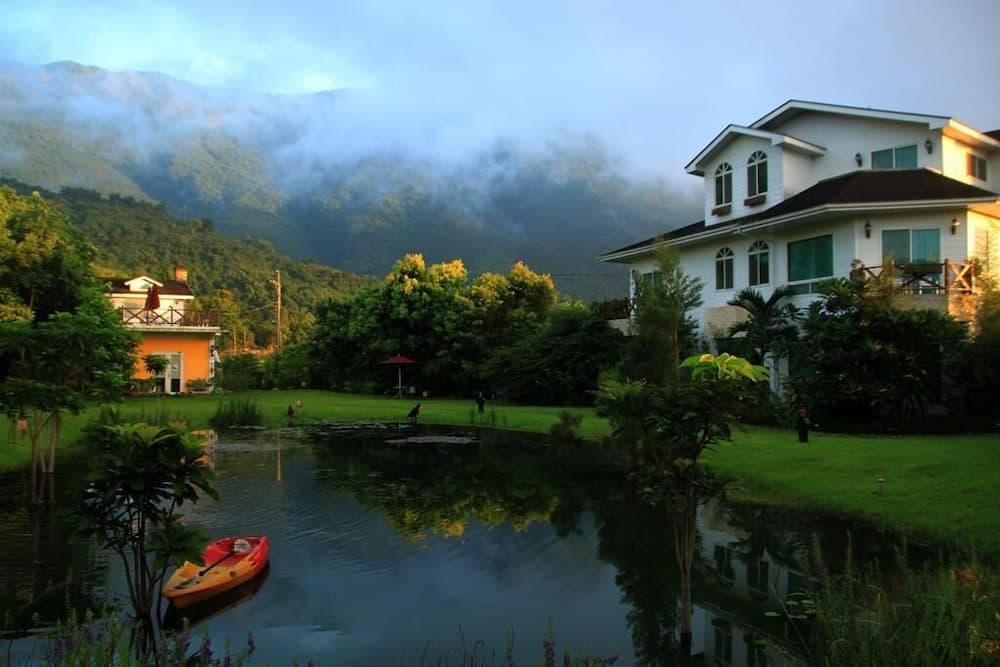 A serene lakeside retreat features a cozy house surrounded by lush greenery and misty mountains, ideal for tranquil vacations with kayaking opportunities.