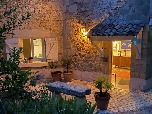 A cozy courtyard illuminated by warm lights features rustic stone walls, potted plants, and an inviting open doorway leading to a charming interior, perfect for travelers seeking a serene and picturesque European retreat.