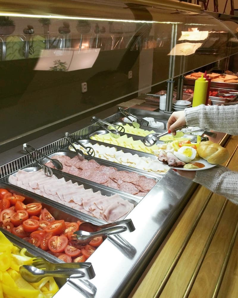 A traveler selects fresh deli items from a vibrant buffet spread in a cozy indoor dining area, perfect for exploring local culinary delights during a vacation.