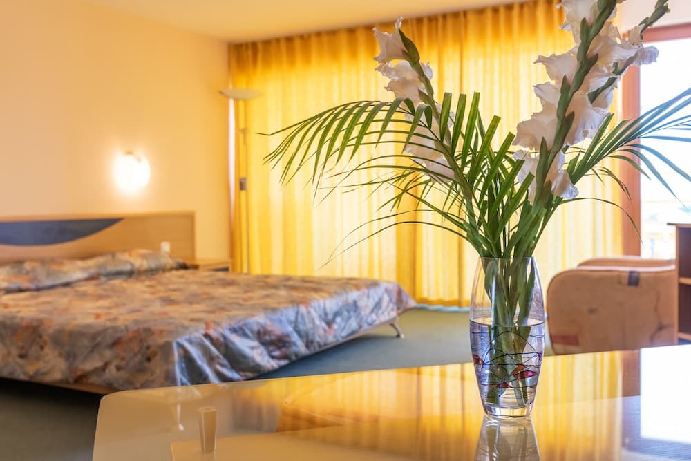 A cozy hotel room bathed in warm light features a floral-patterned bed, yellow curtains, and a vase of white gladioli on a table, ideal for travelers seeking comfort and tranquility.