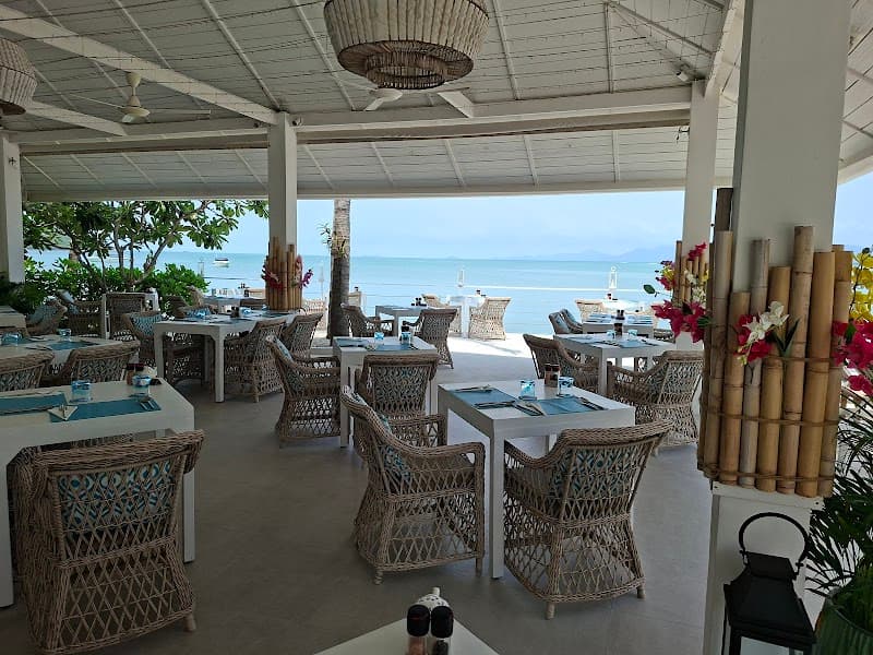 A serene beachside dining area at Tembo Beach Club & Resort in Thailand offers a tranquil atmosphere with wicker furniture, bamboo decor, and stunning ocean views, perfect for travelers seeking relaxation and coastal cuisine.