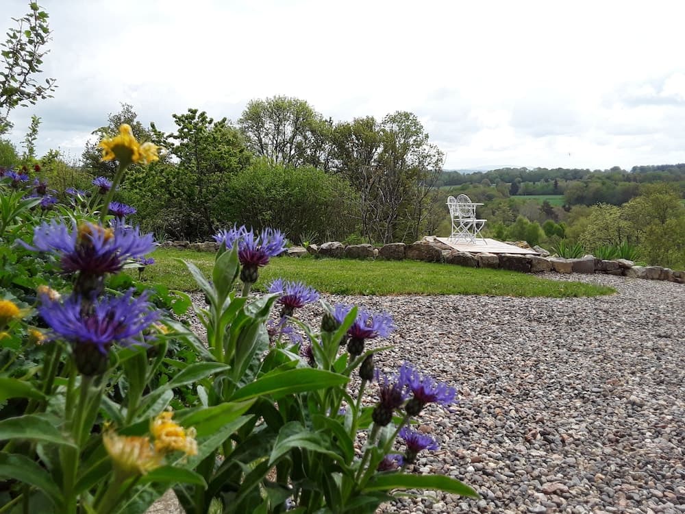 A serene garden path lined with vibrant purple flowers leads to a tranquil stone patio overlooking lush green hills, ideal for a peaceful retreat or nature photography.