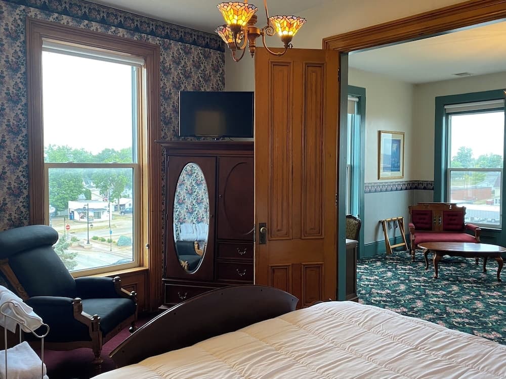 A cozy hotel room features floral wallpaper, a wooden armoire with a mirror, a comfortable bed, and a view of a quiet street through large windows, ideal for travelers seeking relaxation and comfort.